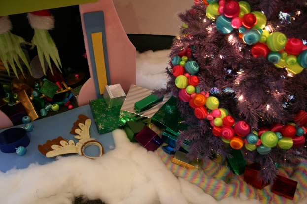 Eine Indoor-Szene mit einem Weihnachtsbaum mit Lichterkette und Dekorationen auf der rechten Seite, Geschenkboxen und Dekorationsgegenständen in der Mitte und einer Hütte mit Dekorationen und Watte in der oberen linken Ecke, mit einer Wand im Hintergrund und Watte und wollener Stoff unten.