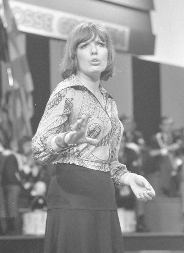 Frau in Schwarz-Weiß-Foto auf einer Bühne stehend, umgeben von einer Gruppe von Menschen mit einer Wand im Hintergrund, weißes Hemd und schwarze Röcke tragend, Haare zu einem Knoten gebunden, ernster Gesichtsausdruck.