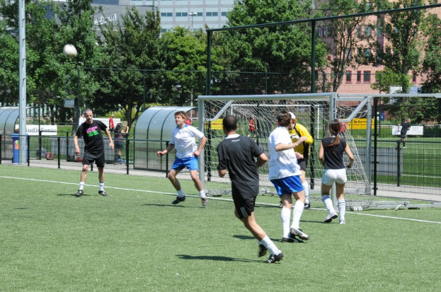 Menschen, die auf einem Feld Fußball spielen, mit einem Zaun und einem Tor in der Mitte, einem Pfosten und einem Ball oben links und Bäumen und Gebäuden im Hintergrund.