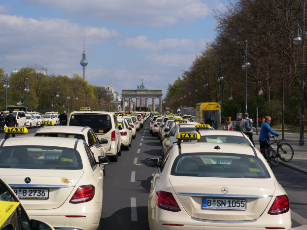 Eine lange Reihe von Taxis steht an der Seite einer belebten Straße in Berlin, Deutschland, mit Fahrzeugen, Fahrradfahrern und Füügängern, flankiert von Laternenmästen, Bäumen und Gebäuden, darunter ein Tor und ein Turm, unter einem bewölktem Himmel.