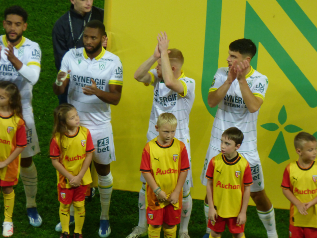 Eine Gruppe junger Menschen in Fußballtrikots steht auf einem Fußballfeld und klatscht feierlich mit einer 'Ligue 1'-Plakette im Hintergrund.