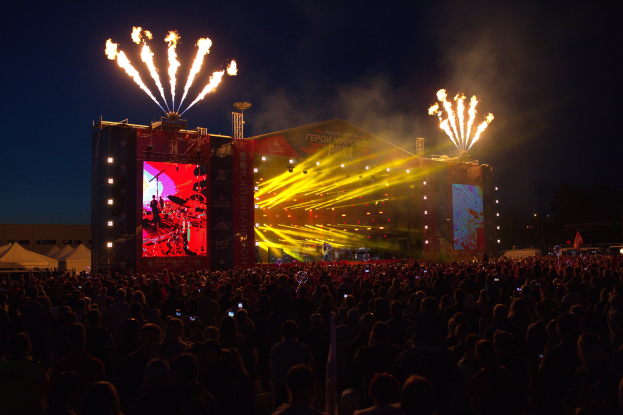Große Menschenmenge auf einem Musikfestival mit einer hell erleuchteten Bühne mit Performern, umgeben von Bannern, Zelten links und Feuerwerk im dunklen Himmel als Hintergrund.