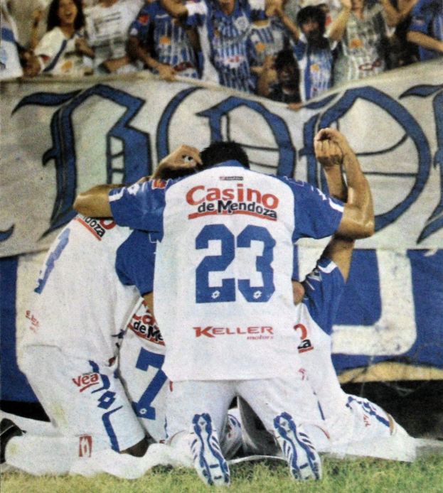 Gruppe von Fußballspielern, die auf dem Boden vor einer jubelnden Menge feiern, mit einem Banner im Hintergrund, auf dem Text zu sehen ist.