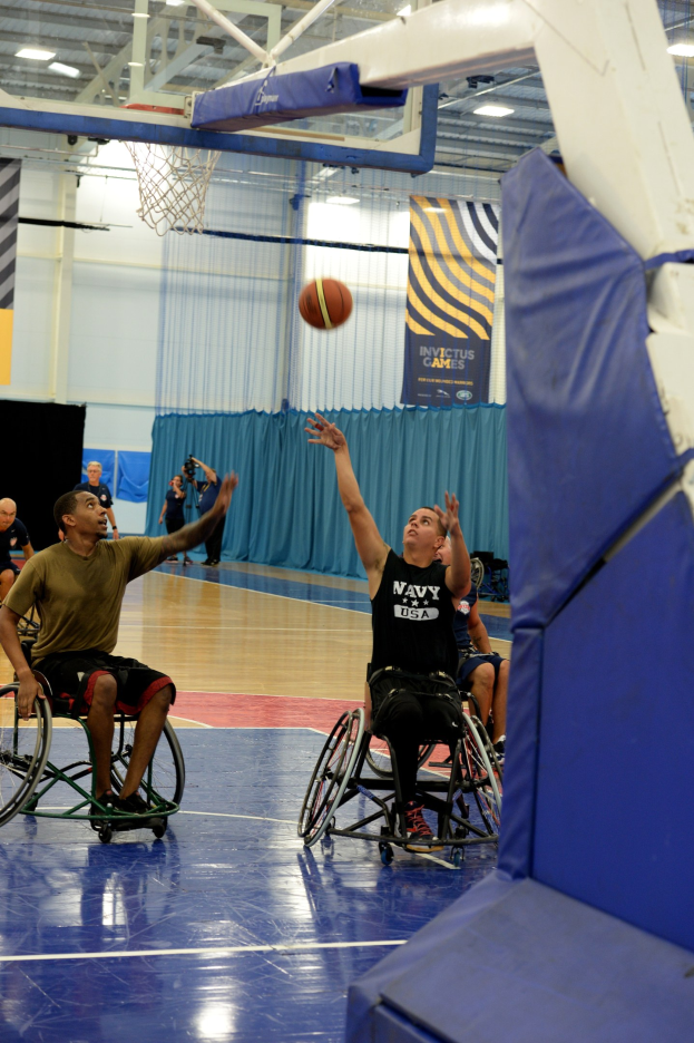 Gruppe von Menschen in Rollstühlen, die Basketball in einer Turnhalle spielen, mit einem Basketballkorb, Vorhängen an der Wand, Bannern mit Text und Deckenbeleuchtung.