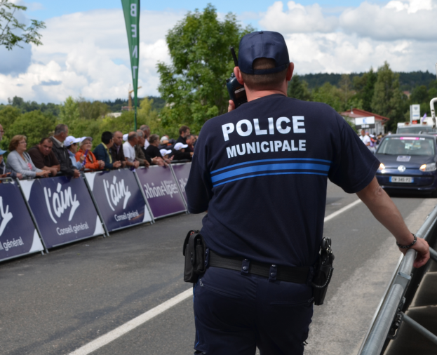 Ein Polizeibeamter in einer Mütze hält ein Funkgerät, während er vor einer Menge mit Schildern, Fahrzeugen, Bäumen, Gebäuden und einem bewölkten Himmel steht.