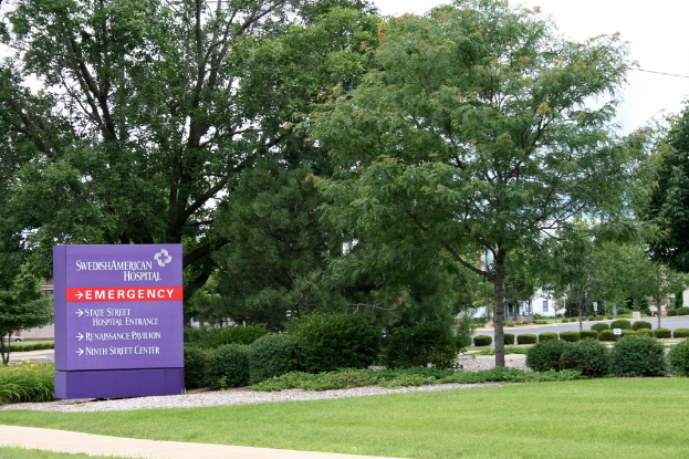 Grass am Boden, ein violettes Schild mit "NOTFALL" hervorgehoben daneben, Bäume und Büsche dahinter, mit Gebäuden und einer Straße im Hintergrund.