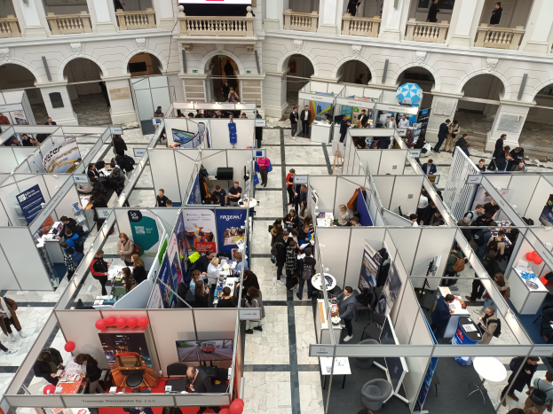 Eine große Gruppe von Menschen versammelt sich in einem Raum mit Ständen, Bannern, Tischen, Stühlen und Ballons, mit einem Gebäude mit Bögen, Säulen, Geländern und einem Bildschirm im Hintergrund.