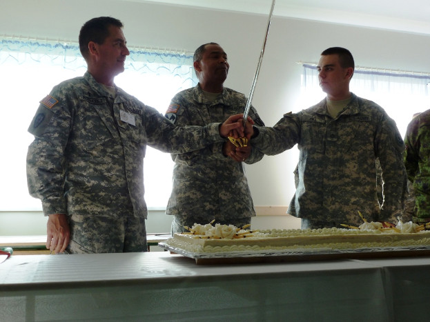 Ein großer Kuchen auf einem Tisch mit mehreren Soldaten davor, drei halten ein großes Messer und eine Wand mit zwei Fenstern dahinter.