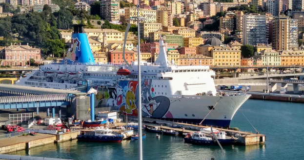 Ein großes Kreuzfahrtschiff liegt nahe einer Stadt mit einer Brücke links, Fahrzeugen auf der Straße und Gebäuden und Bäumen im Hintergrund.