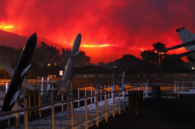 Ein Flugzeug fliegt über eine Start- und Landebahn mit Raketen, umgeben von Zäunen, Mülltonnen, Bäumen, Laternenmasten und Gebäuden, mit einem von den Waldbränden in Kalifornien verqualmten Himmel im Hintergrund.