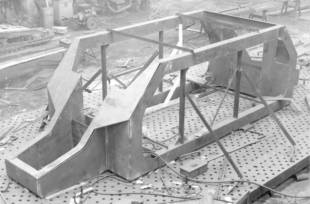 Schwarzes und weißes Foto eines Autos Skeletts auf einem Fabrikboden mit verstreuten Werkzeugen und Maschinen im Hintergrund.