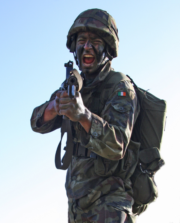 Eine Person in militärischer Uniform mit Abzeichen, Tasche, Helm und einer Waffe.