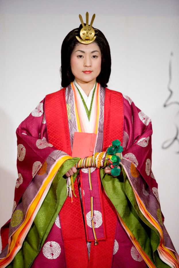 Eine Frau in einem traditionellen japanischen Kimono, das ein leuchtendes rotes und grünes Kleid mit aufwändigen Mustern trägt, steht selbstbewusst auf einem Laufsteg, ihr Haar zu einem ordentlichen Knoten gebunden.