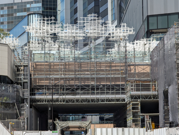 Großes Gebäude mit Gerüst in einer städtischen Umgebung umgeben von Bäumen auf der linken Seite und einer Straße unten.