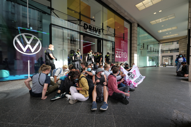 Eine Gruppe von Menschen sitzt vor einer Volkswagen-Vertretung mit Glaswänden und einem Logo auf der linken Seite auf dem Boden, einige halten ein Banner mit Text, unter Deckenleuchten, scheinbar protestierend.