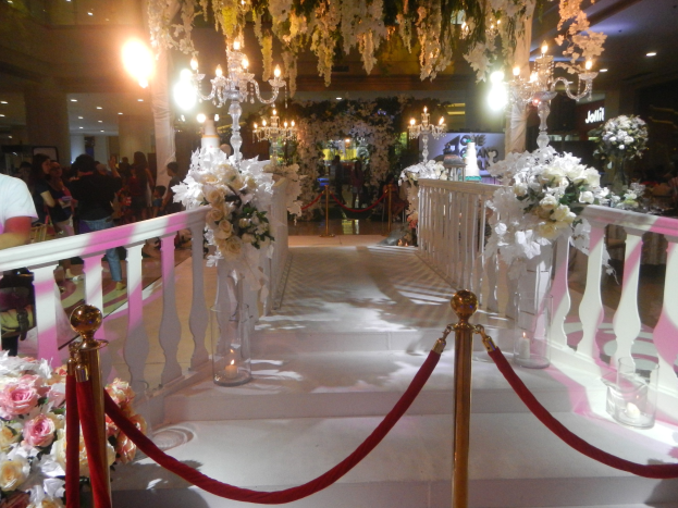 Eine Hochzeitszeremonie in einem Einkaufszentrum mit weißem Teppich, dekorativen Geländern, Blumenschmuck und Deckenbeleuchtung, bei der zahlreiche Gäste anwesend sind.