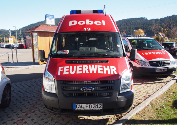 Ein Rettungswagen auf einem Parkplatz mit anderen Autos, Gras, Pfählen, Brettern, einer Hütte, Bäumen, Hügeln und einem klaren blauen Himmel.