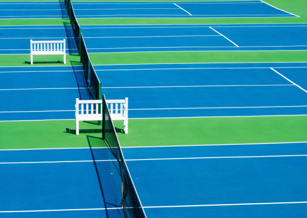 Ein blau-grüner Tennisplatz mit zwei weißen Bänken und einem Netz in der Mitte, umgeben von saftigem Grün.