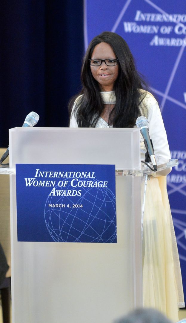 Eine Frau in einem weißen Kleid und Brille steht an einem Podium mit zwei Mikrofonen und einer Tafel, während sie vor einer blauen Fahne mit der Aufschrift "International Women of Courage Awards 2014" eine Menge anspricht.