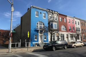 Eine Reihe bunter Häuser an einer Straßenecke in Baltimore, Maryland, mit parkenden Autos, Bäumen, Pfählen, Brettern und einem Zaun, der die Straße säumt, unter einem bewölkten Himmel.