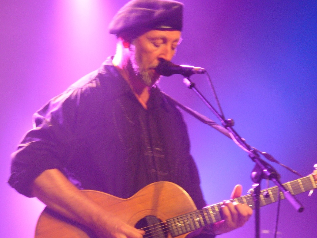Eine Person auf einer Bühne mit einer Mütze, die eine Gitarre hält und spielt, während sie ein Lied singt.