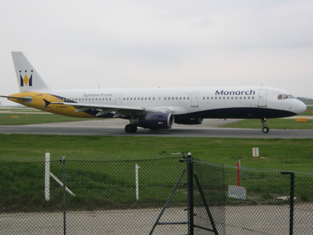 Ein Monarch Airlines Airbus A320-200 auf einer Rollbahn mit Gras, einem Zaun, Bäumen und einem klaren blauen Himmel.