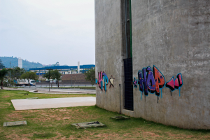 Betongebäude mit Graffiti, umgeben von Gras, Bäumen, Strommasten, Fahrzeugen, anderen Gebäuden, Hügeln und einem klaren blauen Himmel.