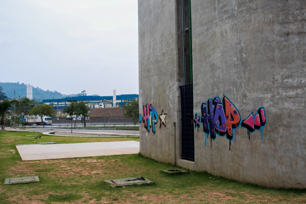 Betongebäude mit Graffiti, umgeben von Gras, Bäumen, Strommasten, Fahrzeugen, anderen Gebäuden, Hügeln und einem klaren blauen Himmel.