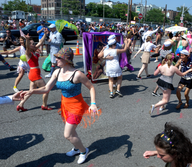 Eine Gruppe von Menschen, die eine Straße entlang marschieren, einige mit Mützen und Schirmen, andere tanzend, mit einem Banner, Verkehrskegeln, Fahrzeugen, Laternen, Schildern, Bäumen, Gebäuden und einem klaren blauen Himmel im Hintergrund.