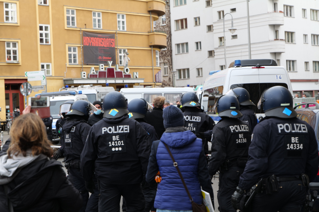 Eine Gruppe von Polizisten steht vor einer Menge von Menschen, die Helme und Jacken tragen, während einer Demonstration in Berlin, Deutschland, mit Fahrzeugen, Gebäuden, Laternenpfählen und einem Banner mit Text im Hintergrund, und einer Person mit einer Kamera.