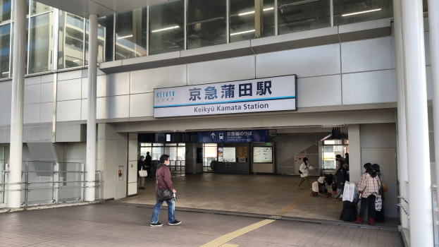 Menschen mit Gepäck durch einen Bahnhof gehen, eine Abfahrtsanzeige, Säulen, Geländer, Glaswände und Deckenleuchten.