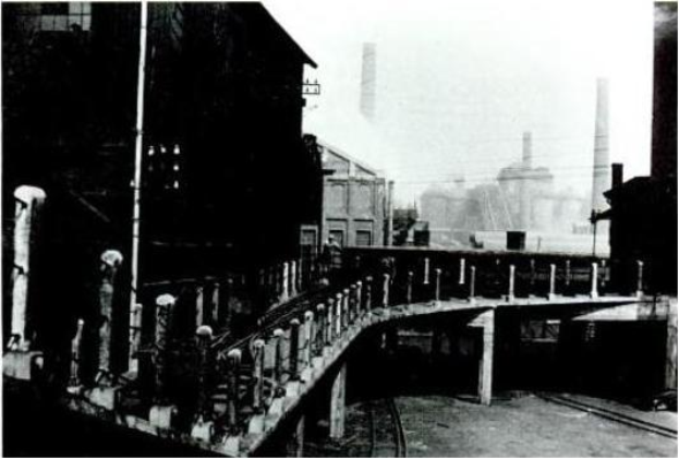 Ein Schwarz-Weiß-Foto einer Brücke über eine Bahnschiene, mit Gebäuden auf beiden Seiten und einer Fabrik im Hintergrund, mit Geländern und Pfählen entlang der Seiten der Überführung.