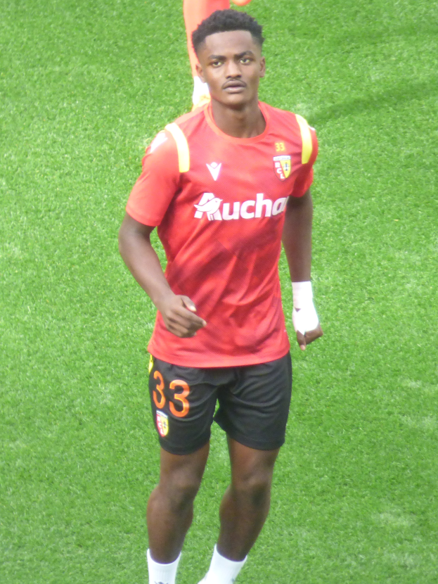 Emmanuel Adebayor, ein Mann in einem roten Trikot und schwarzen Shorts, läuft auf einem Fußballfeld mit zwei Personen im Hintergrund.
