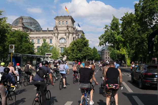 Eine Gruppe von Menschen, die auf Fahrrädern eine von Bäumen gesäumte Straße in Berlin, Deutschland, entlangfährt, mit Gebäuden auf beiden Seiten, einem Bushaltestellenschild auf der rechten Seite und einer Flagge, die auf einem der Gebäude weht, unter einem bewölkten Himmel.