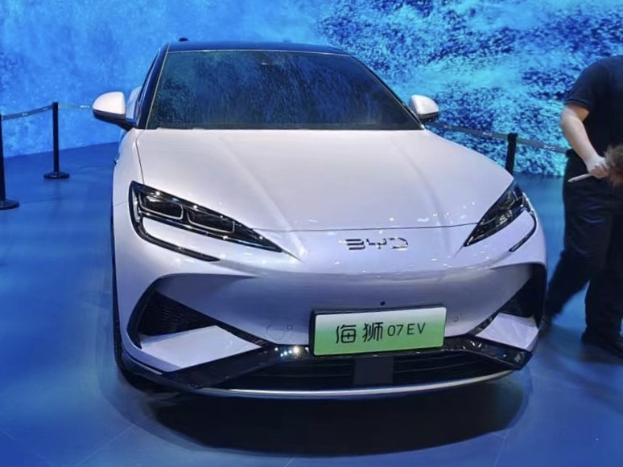 Weißes Auto auf einer Automesse mit einer Person rechts daneben, die ein Objekt hält, einem Bildschirm im Hintergrund und Absperrpollern mit Seilen links.