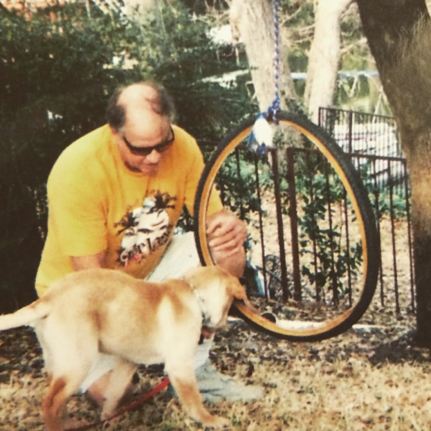 Ein älterer Mann in einem gelben T-Shirt und Brille steht auf dem Boden mit einem Hund vor ihm, ein Reifen an einen Baum mit einem Seil im Hintergrund, zusammen mit einem Zaun und mehr Bäumen.