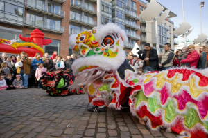 Ein lebendiges chinesisches Neujahrsfest in Amsterdam mit einer Löwen-Tanz-Show vor einer Menschenmenge, einige halten Kameras, vor dem Hintergrund von Gebäuden, Laternenmasten und einem klaren blauen Himmel.