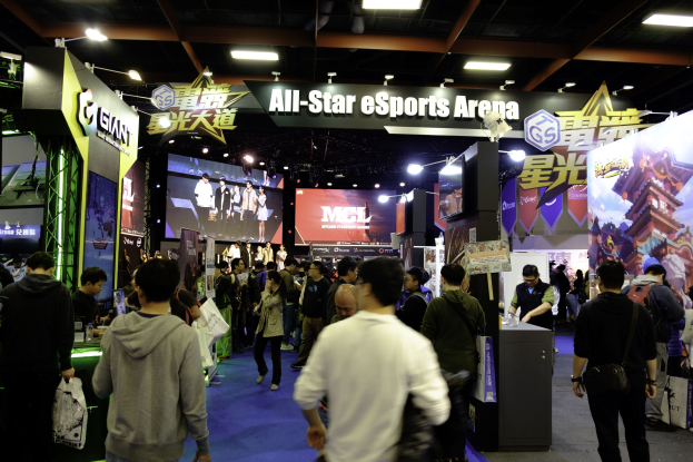 Eine große Gruppe von Menschen steht vor einem Stand in einer All-Star-E-Sports-Arena, einige halten Taschen, andere umringen Tische mit verschiedenen Gegenständen, während Banner und beleuchtete Bildschirme zur lebhaften Atmosphäre beitragen.
