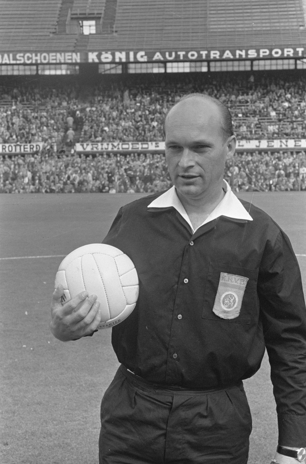 Schwarz-weiß-Foto eines Schiedsrichters, der einen Fußball auf einem Feld hält, mit Zuschauern und Tafeln im Hintergrund.