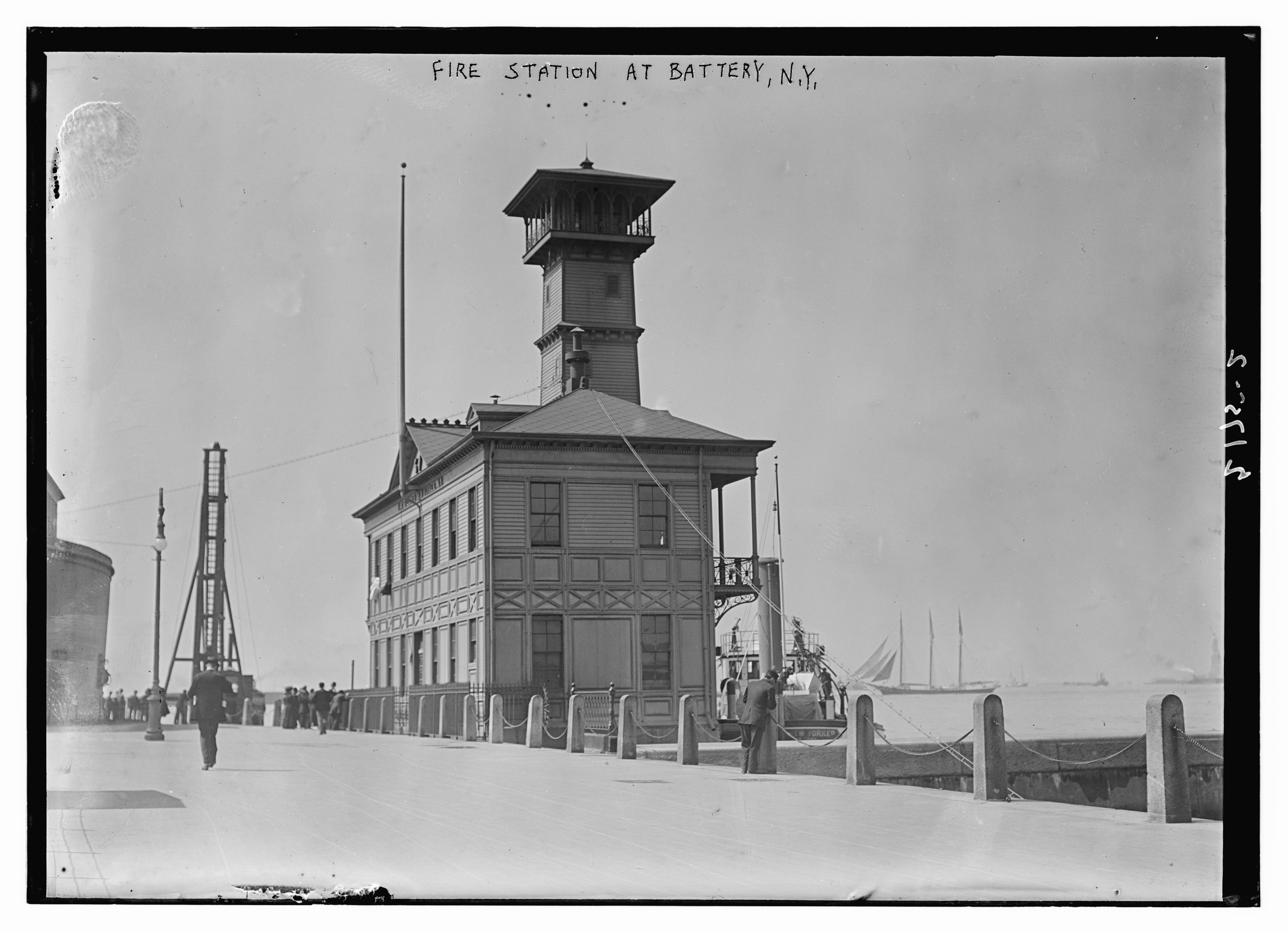 Ein Schwarz-Weiß-Foto einer Feuerwache an der Battery, New York, mit einer Gruppe von Menschen auf dem Boden, Pfählen, einem Zaun, Booten auf dem Wasser und einem bewölkten Himmel.