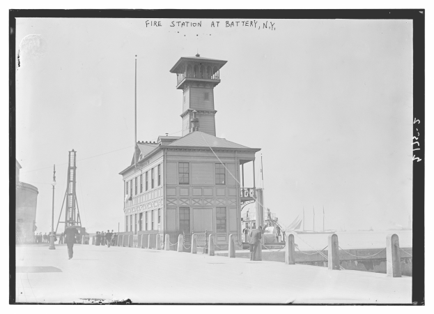 Ein Schwarz-Weiß-Foto einer Feuerwache an der Battery, New York, mit einer Gruppe von Menschen auf dem Boden, Pfählen, einem Zaun, Booten auf dem Wasser und einem bewölkten Himmel.