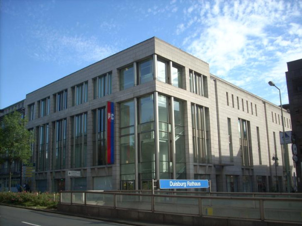 Ein großes Gebäude mit zahlreichen Fenstern, das als Hauptsitz der Deutschen Bank dient, umgeben von Straßenmasten, Straßenlaternen, Namensschildern, Kraftfahrzeugen, Büschen, Bäumen und einem bewölkten Himmel.