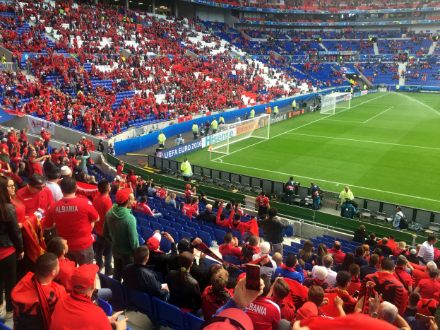 Eine große Menschenmenge in einem Stadion schaut einem Fussballspiel zu, mit Menschen, die auf Stühlen sitzen und stehen, Schildern, einem Torpfosten und Deckenlichtern.