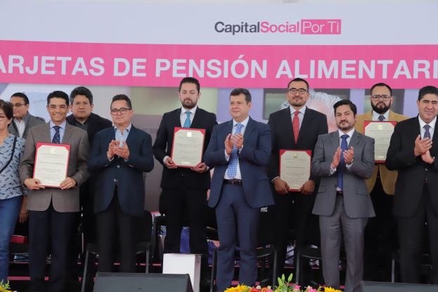 Eine Gruppe von Menschen auf einer Bühne, die klatschen und Zertifikate halten, mit Stühlen in einem Halbkreis und einem Banner mit der Aufschrift "Capital Social for Ti" im Hintergrund sowie Blumensträußen und Rednern unten.