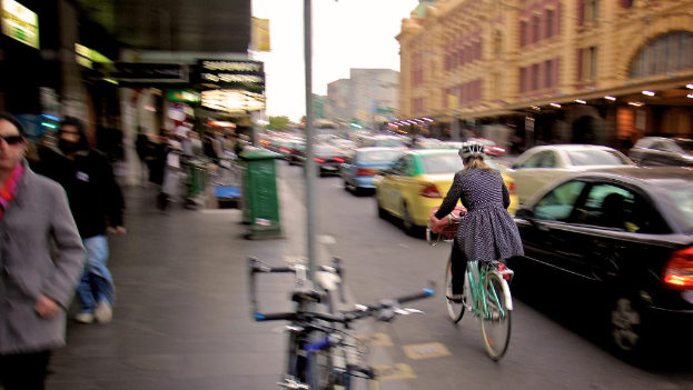 Ein M√∂dchen mit Helm f√§hrt ein Fahrrad auf einer vielbefahrenen Stra√Ÿe mit vielen Fahrzeugen, Geb√§uden auf der rechten Seite, Fussg√§ngern auf der linken Seite, einem abgestellten Fahrrad auf dem Gehweg und Gesch√§ften mit Lichtern.