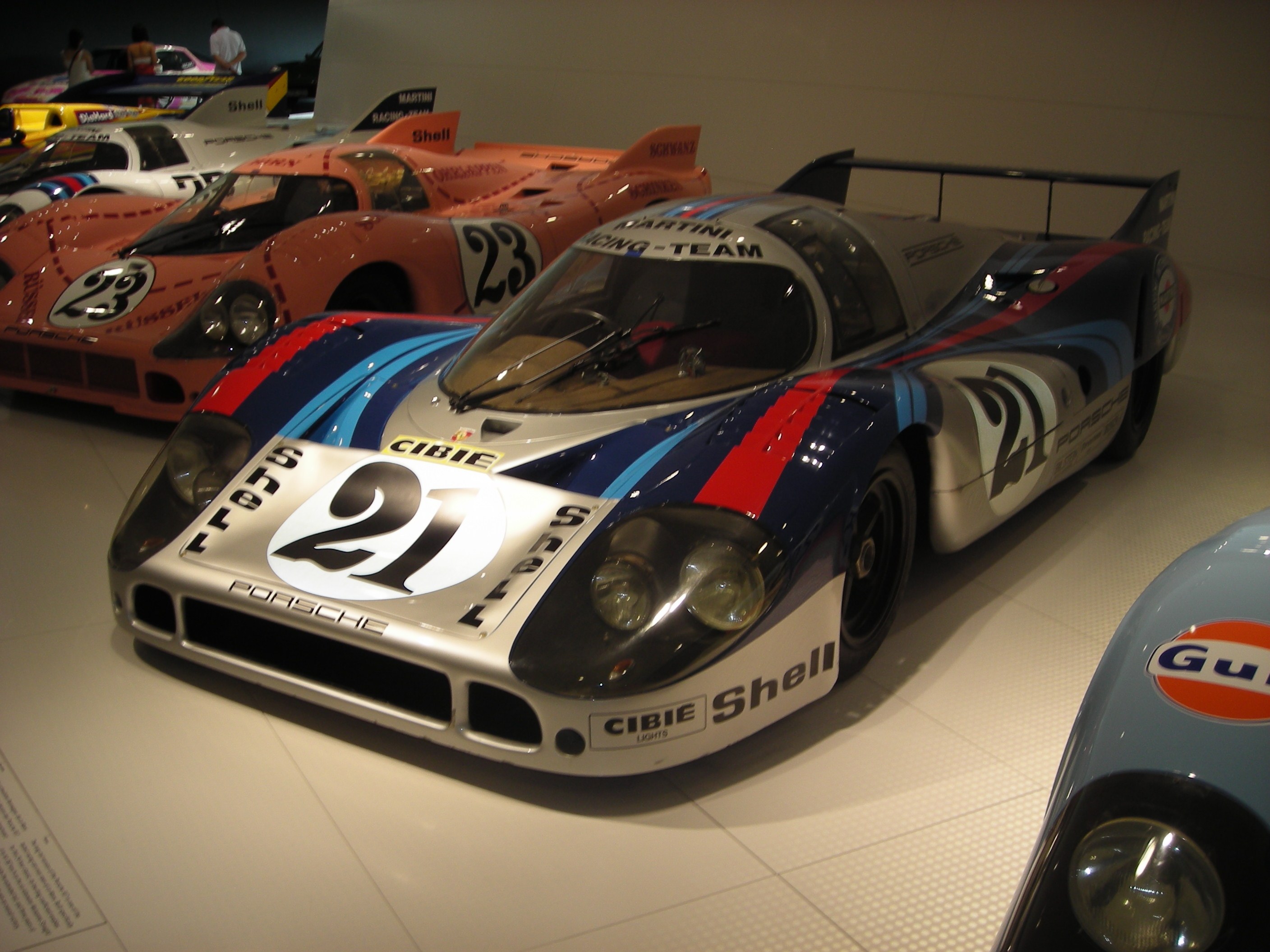 Ein Porsche 917K Rennwagen auf einer Museumswand, umgeben von anderen Sportwagen, mit Menschen in der Nähe und einer Wand im Hintergrund.
