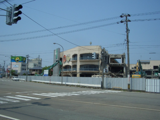 Eine Stadtstraße mit fahrenden Fahrzeugen, Strommasten, Laternenmasten und einer Ampel auf der linken Seite, mit Gebäuden, Bäumen und dem Himmel im Hintergrund und einem abbruchreifen Gebäude im Vordergrund.