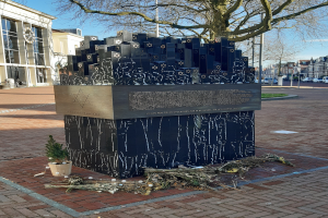 Ein Denkmal für Holocaust-Opfer steht in einem Stadtplatz, mit einem Baum davor, einem Topfgewächs, verstreuten trockenen Blättern, umgeben von Gebäuden, Fahrzeugen, Pfählen, Bäumen und dem Himmel im Hintergrund.