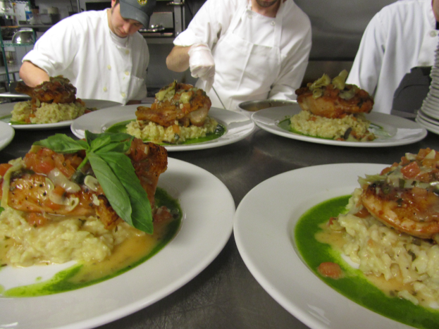 Drei Köche in einer Küche mit Mützen und Handschuhen bereiten Essen auf Tellern zu, mit Regalen im Hintergrund.