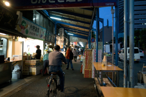 Ein Gehweg mit Geschäften auf der linken Seite und Kisten, einem Tisch, einer Straße mit Autos, Bäumen und einem Gebäude auf der rechten Seite, mit Menschen, die gehen und einer Person, die ein Fahrrad fährt.